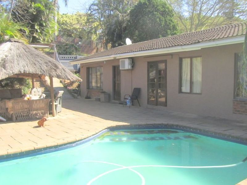 Rear exterior view featuring a swimming pool, paved patio area, and a thatched lapa structure set against the house facade.