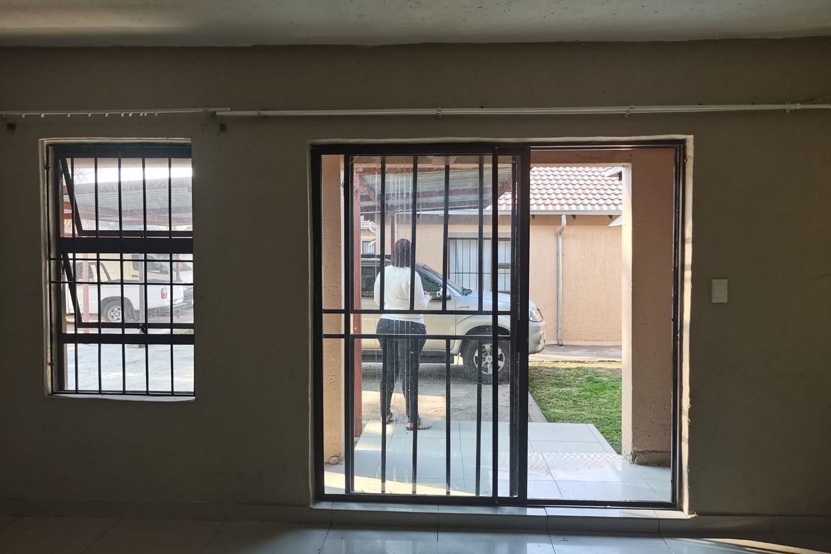 An empty living area featuring tiled flooring, security bars on windows and sliding doors, with direct access to the exterior.