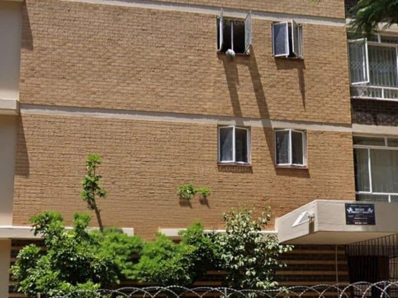 Exterior view of a multi-story brick apartment building featuring security fencing, razor wire, and an entrance canopy.