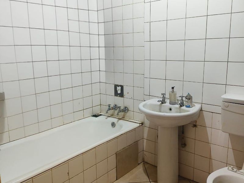 A dated bathroom featuring a standard bathtub, pedestal basin, and toilet, finished with white wall tiles and neutral floor tiles.