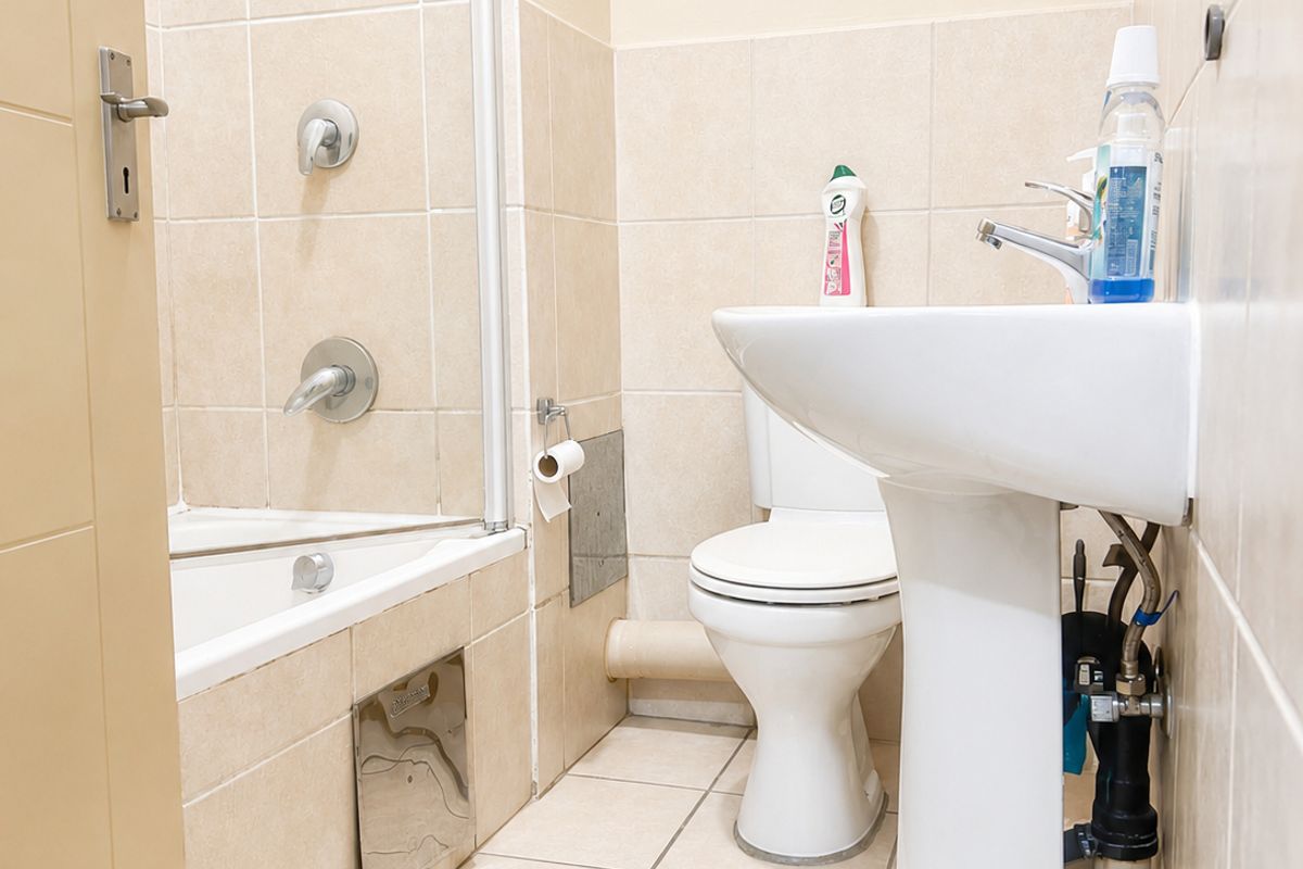 A functional bathroom featuring a pedestal basin, toilet, and a bathtub with a glass shower screen, finished with neutral wall and floor tiles.