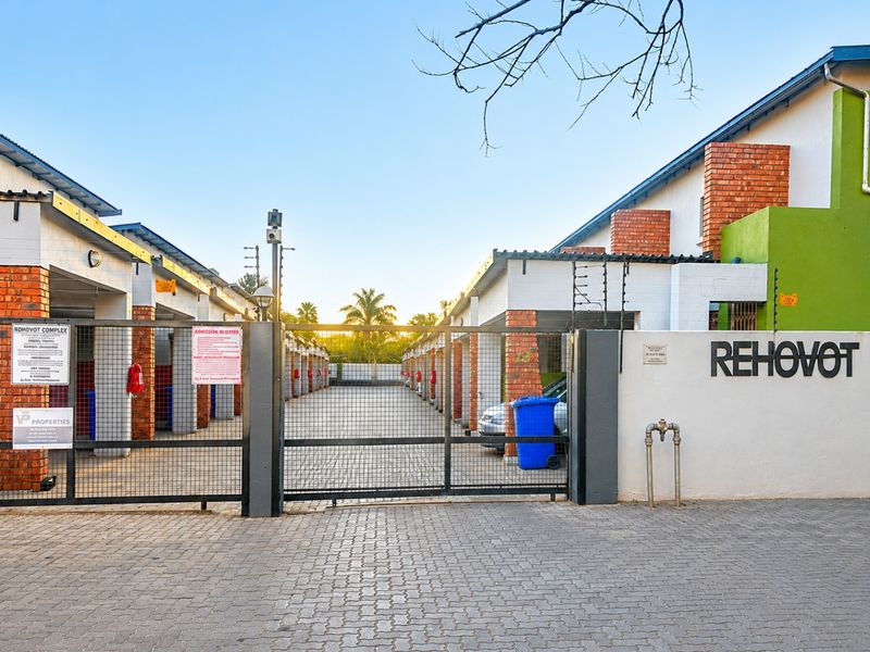 Front entrance view of the Rehovot complex, featuring secure gated access, brick-paved driveway, and perimeter electric fencing.