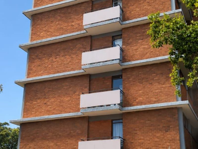 A multi-story apartment building featuring a red brick facade, stacked concrete balconies with metal railings, and secure perimeter fencing.
