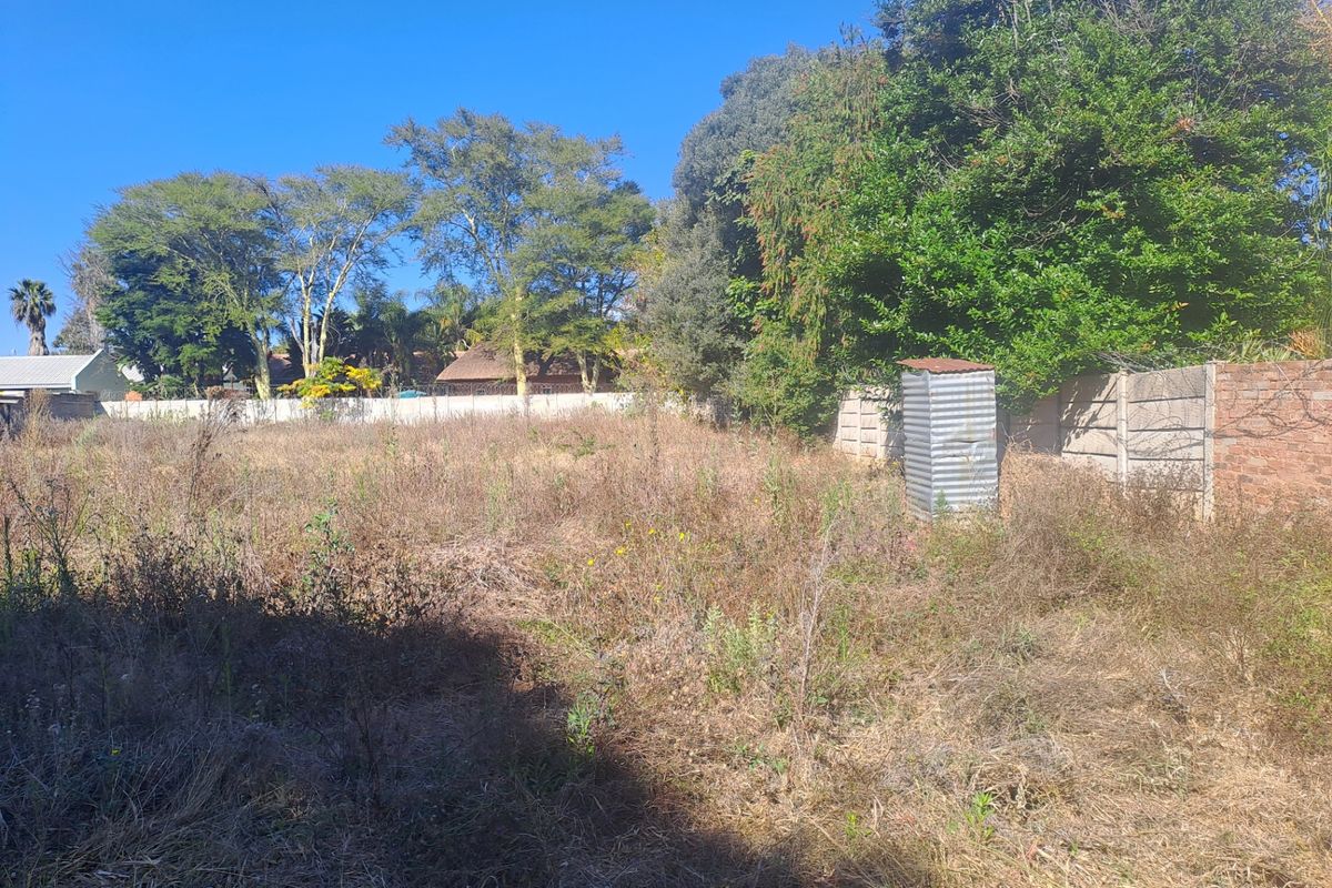 A vacant residential plot featuring overgrown grass, mature trees, perimeter walling, and a small, weathered corrugated metal utility structure.