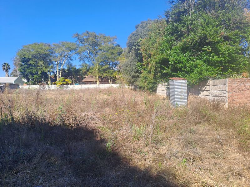 A vacant residential plot featuring overgrown grass, mature trees, perimeter walling, and a small, weathered corrugated metal utility structure.