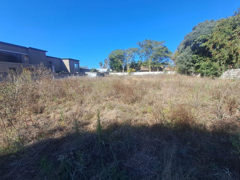 A vacant residential plot featuring overgrown grass and shrubs, bordered by existing boundary walls and mature trees in a suburban setting.