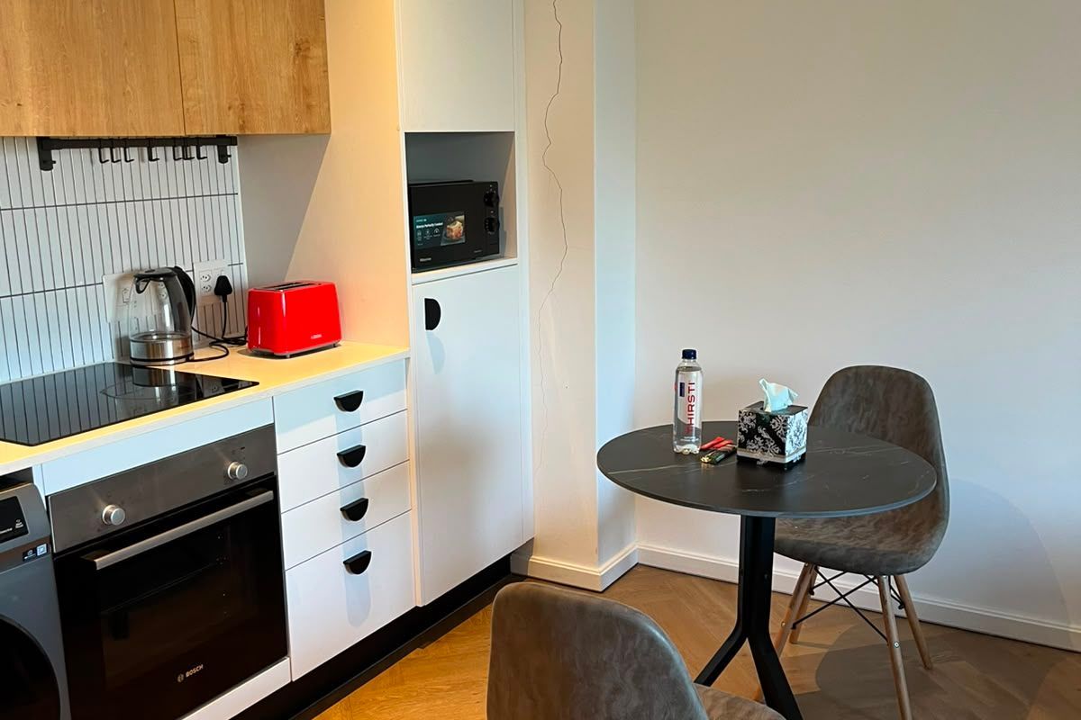 Compact modern kitchen featuring light wood-tone cabinetry, integrated appliances, herringbone timber flooring, and a tiled splashback, with a small adjacent dining area.