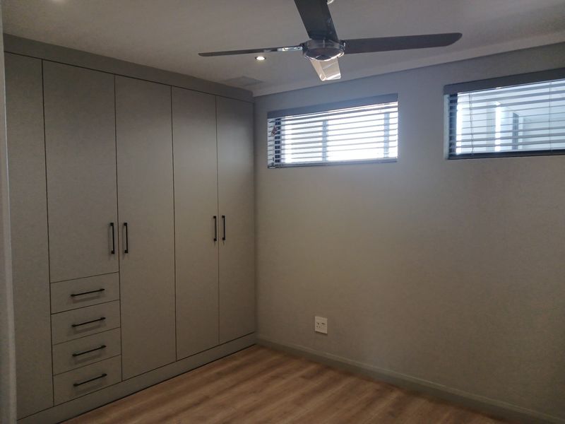 A modern bedroom featuring neutral grey built-in cabinetry, wood-look flooring, a ceiling fan, and horizontal window blinds.