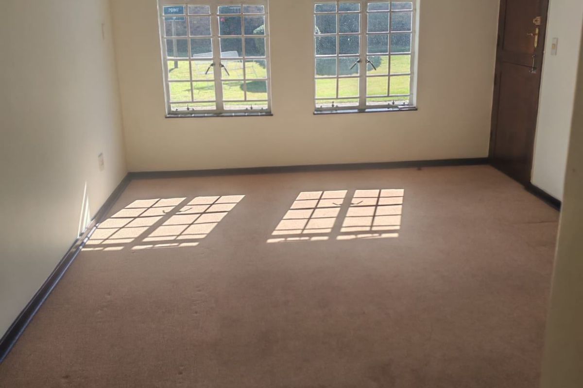 A bright, empty bedroom featuring neutral walls, carpeted flooring, and large windows that provide natural light and views of the exterior grounds.