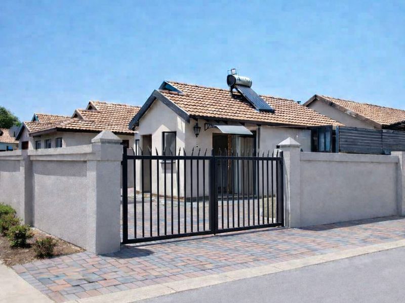 Front view of a residential house featuring a paved driveway, secure perimeter wall with metal gate, and a roof-mounted solar water heater.