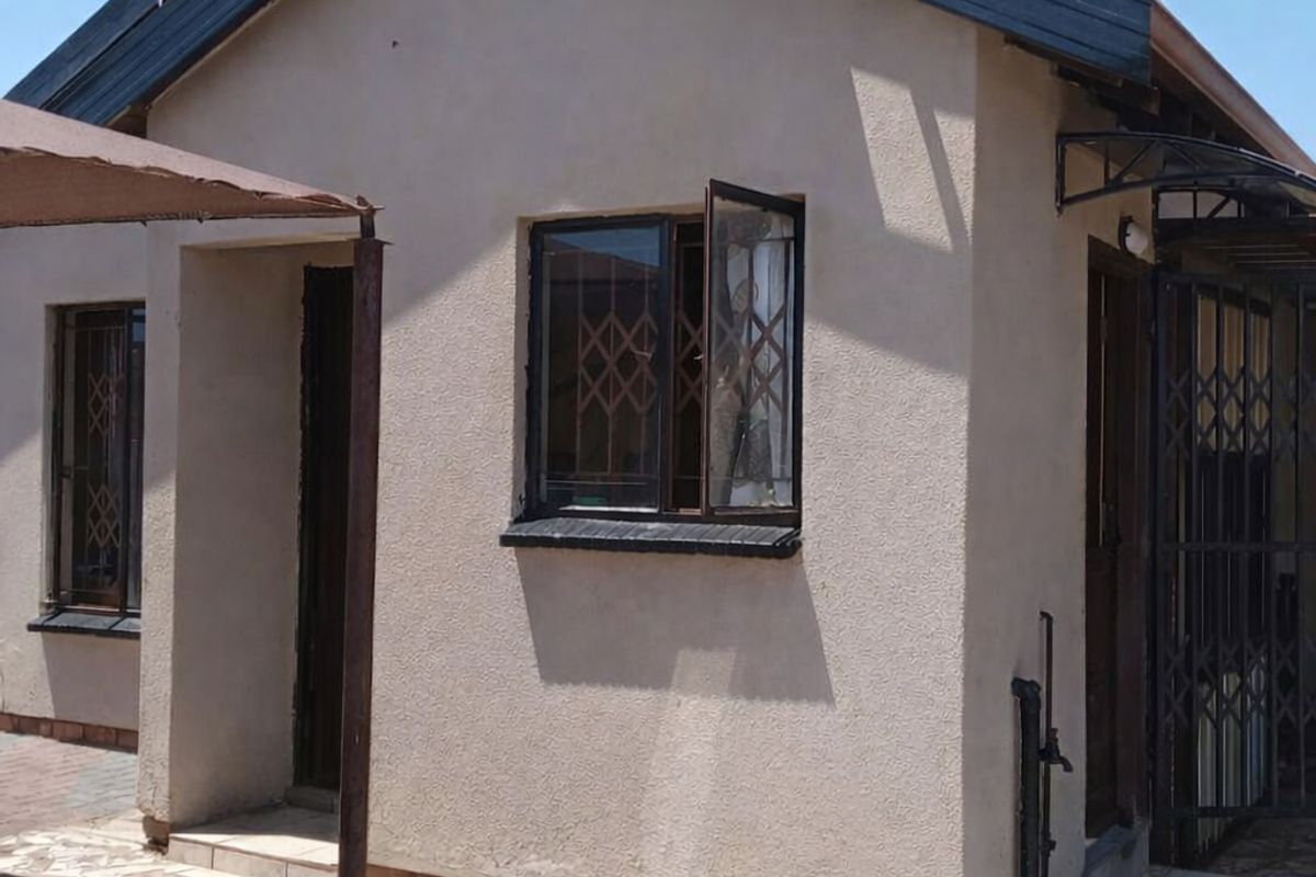 Exterior view of a single-story house featuring a pitched roof, security gates on windows, and paved surrounding area.