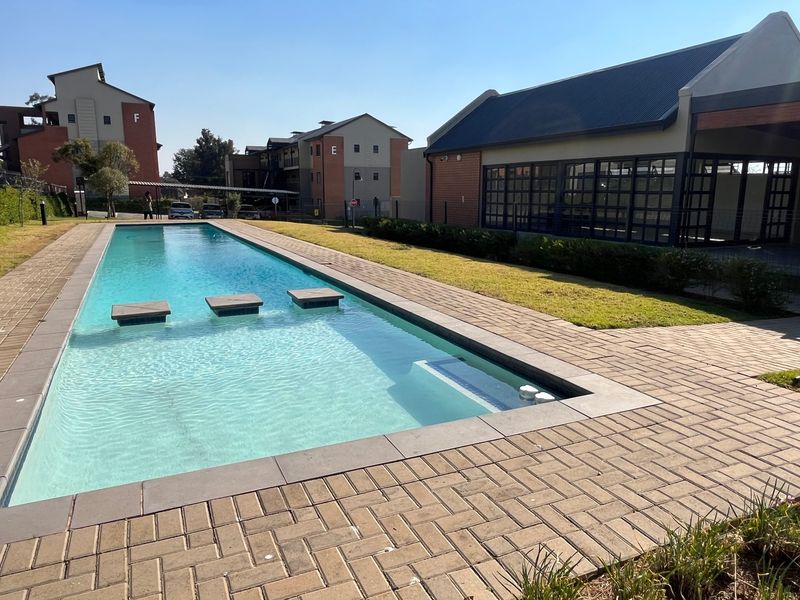 A well-maintained communal swimming pool area featuring paved surrounds, landscaped lawns, and adjacent clubhouse facilities within a modern apartment complex.