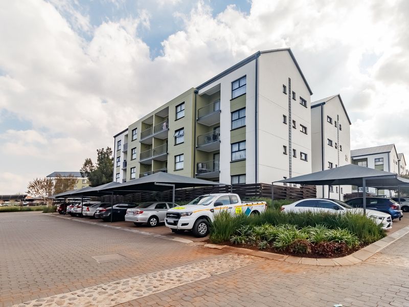 Exterior view of a modern multi-story apartment complex featuring paved parking areas with shade-cloth carports and landscaped garden beds.