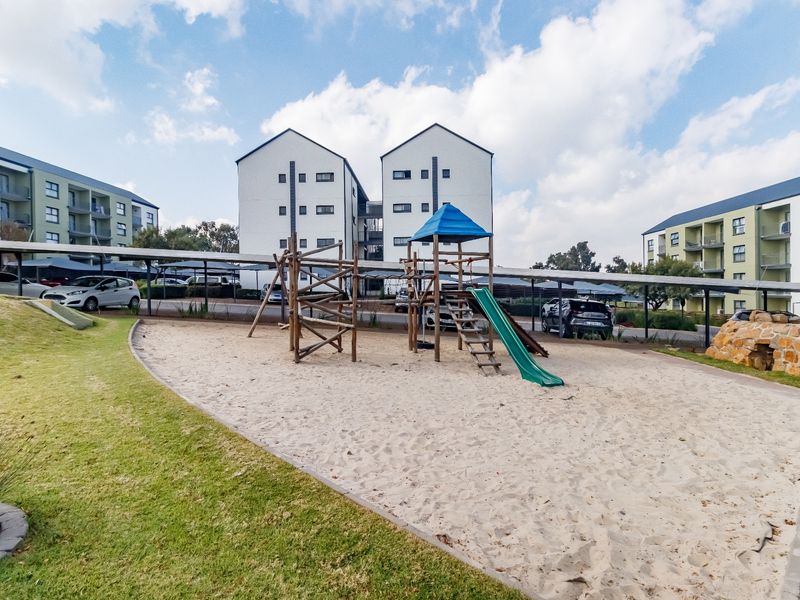 Communal outdoor area featuring a sand-based playground, landscaped lawn, and covered parking bays within the apartment complex grounds.