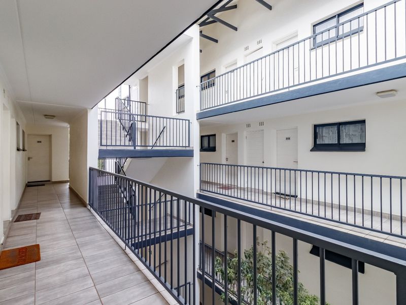 Open-air apartment complex hallway featuring multi-level walkways, metal safety railings, and tiled flooring providing access to various residential units.