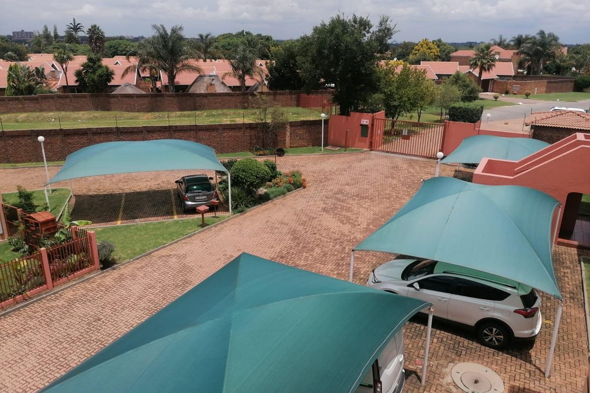 Elevated view of the apartment complex parking area featuring paved driveway, landscaped garden beds, and multiple green shade-cloth carports.
