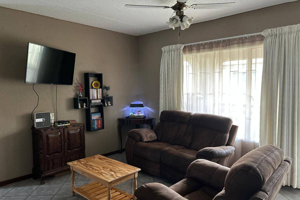 A comfortable living room featuring tiled flooring, a ceiling fan, and large windows with curtains, providing a functional and well-lit space.