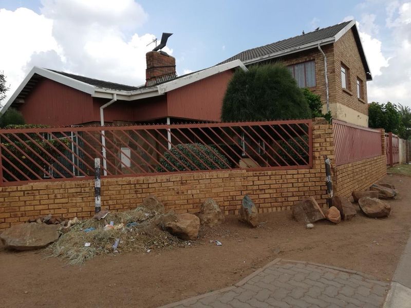 Street view of a multi-level brick house featuring a tiled roof, chimney, and perimeter wall with metal security fencing.