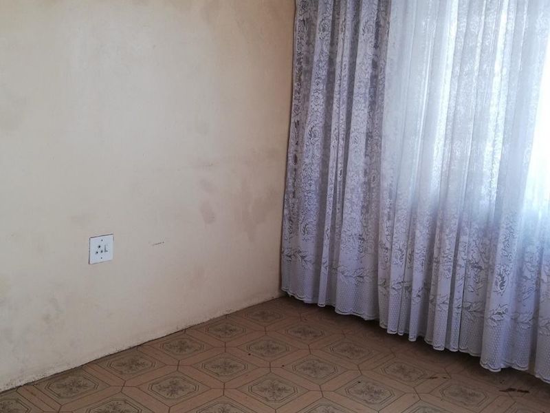 A bedroom featuring patterned vinyl flooring and a window with curtains, showing signs of wear and requiring cosmetic updates.