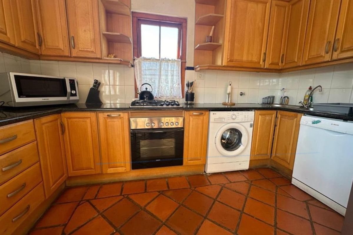 A functional kitchen featuring wooden cabinetry, granite countertops, a gas hob, built-in oven, and terracotta floor tiles, offering a classic, dated aesthetic.