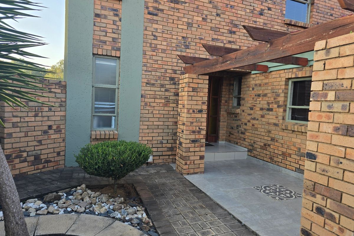 A rear exterior view of the townhouse featuring a brick facade, a paved patio area with a timber pergola, and a landscaped garden section.