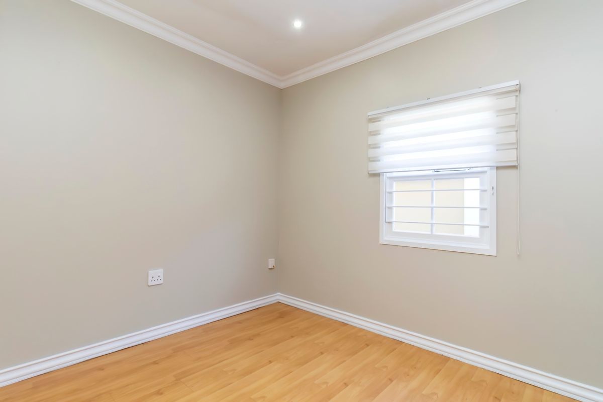 A clean, empty bedroom featuring light-toned laminate flooring, neutral walls, decorative cornice, and a window fitted with a modern roller blind.