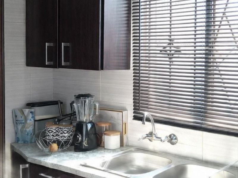 A functional kitchen area featuring dark wood cabinetry, a double stainless steel sink, stone-look countertops, and horizontal window blinds.