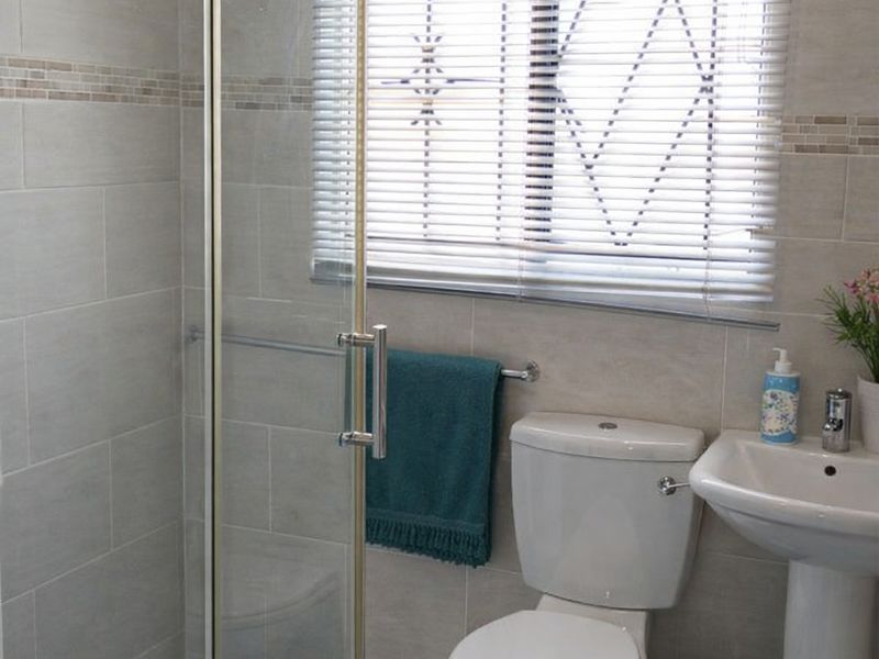 A modern bathroom featuring a glass-enclosed shower, pedestal basin, and toilet, finished with neutral wall tiles and a window with fitted blinds.