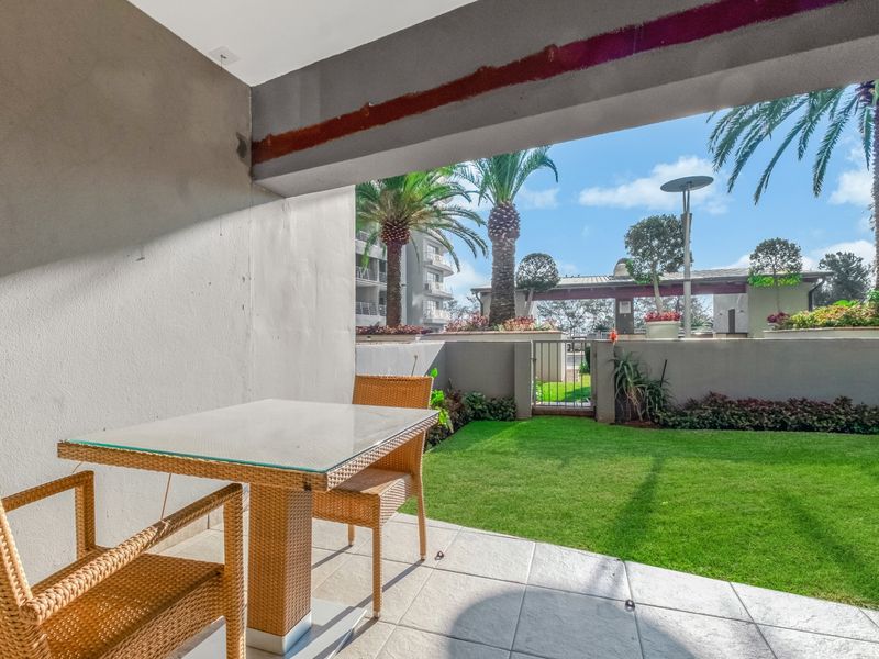 A covered patio area featuring tiled flooring, overlooking a private garden space with landscaped greenery and palm trees.