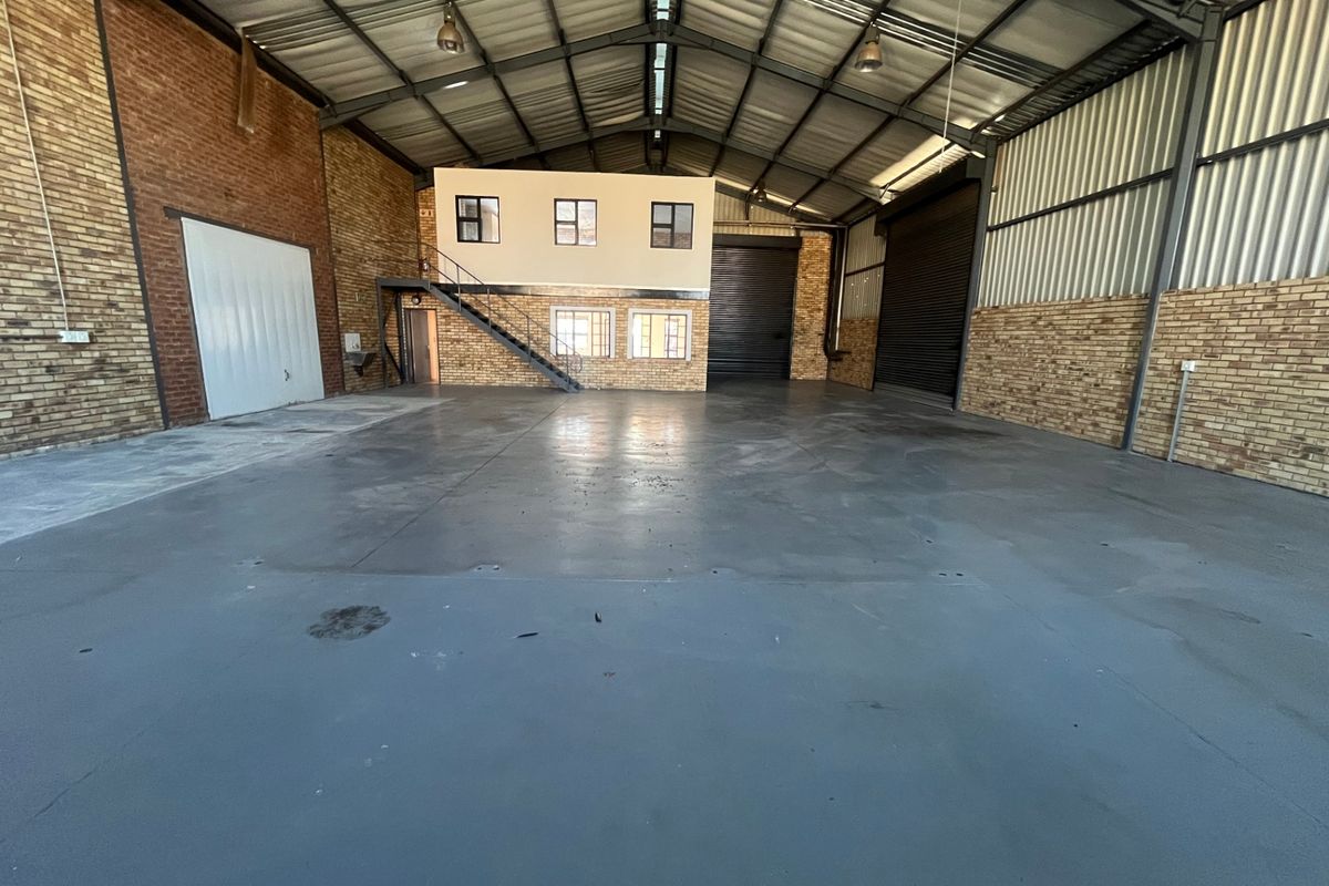 Spacious industrial warehouse featuring high ceilings, exposed brick walls, a mezzanine office level with staircase, and large roller shutter doors for easy access.
