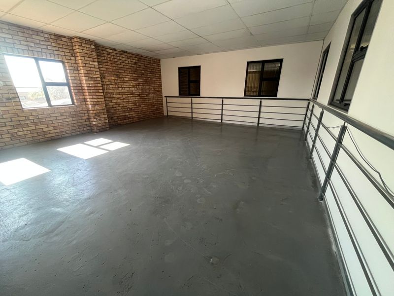 An open-plan industrial mezzanine space featuring exposed brickwork, polished concrete flooring, suspended ceiling tiles, and black-framed windows with metal safety railings.