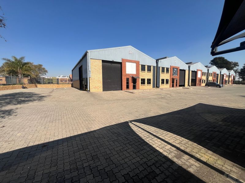 Exterior view of a multi-unit industrial complex featuring brick and metal cladding, roller shutter doors, and a paved parking area.