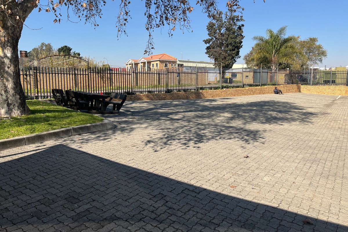 A paved outdoor area featuring a perimeter security fence, brick retaining wall, and established trees, suitable for parking or outdoor utility use.