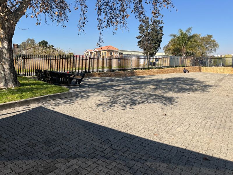 A paved outdoor area featuring a perimeter security fence, brick retaining wall, and established trees, suitable for parking or outdoor utility use.