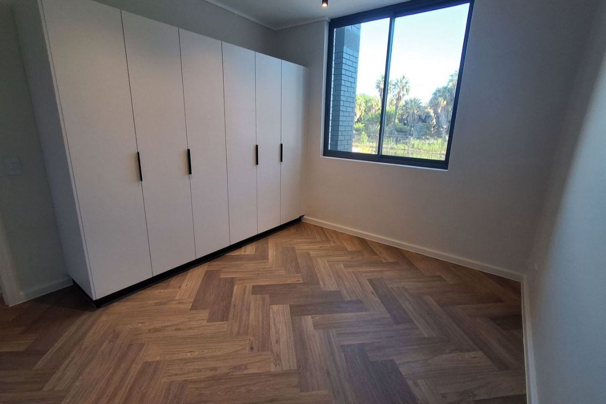 A bright, modern bedroom featuring high-quality herringbone timber flooring, built-in cupboards, and large windows with a pleasant outdoor view.