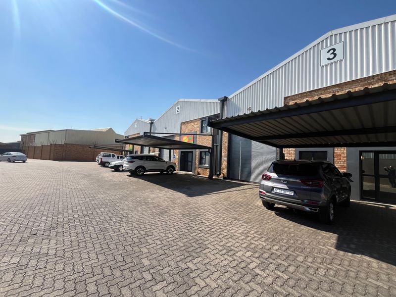 Exterior view of an industrial complex featuring multiple units with brick facades, corrugated metal roofing, and individual covered parking bays.