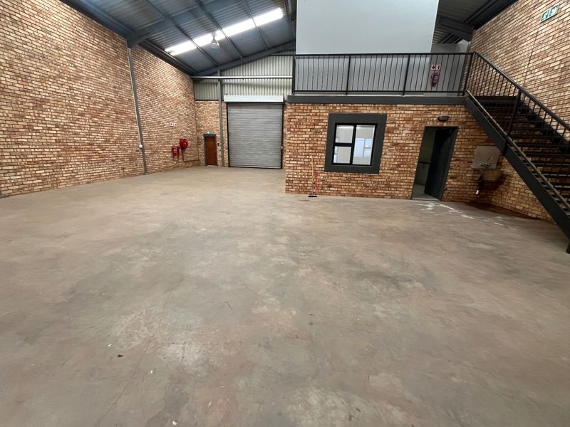 Spacious industrial warehouse unit featuring exposed brick walls, high ceilings, a roller shutter door, mezzanine level with metal staircase, and concrete flooring.