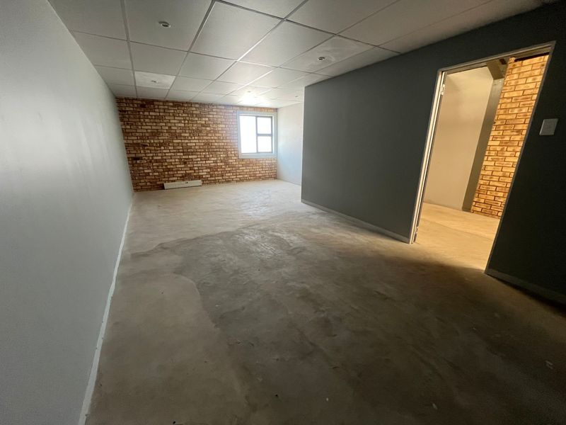An empty industrial office space featuring exposed brick accent walls, a suspended ceiling grid, and concrete flooring with an adjacent room access.