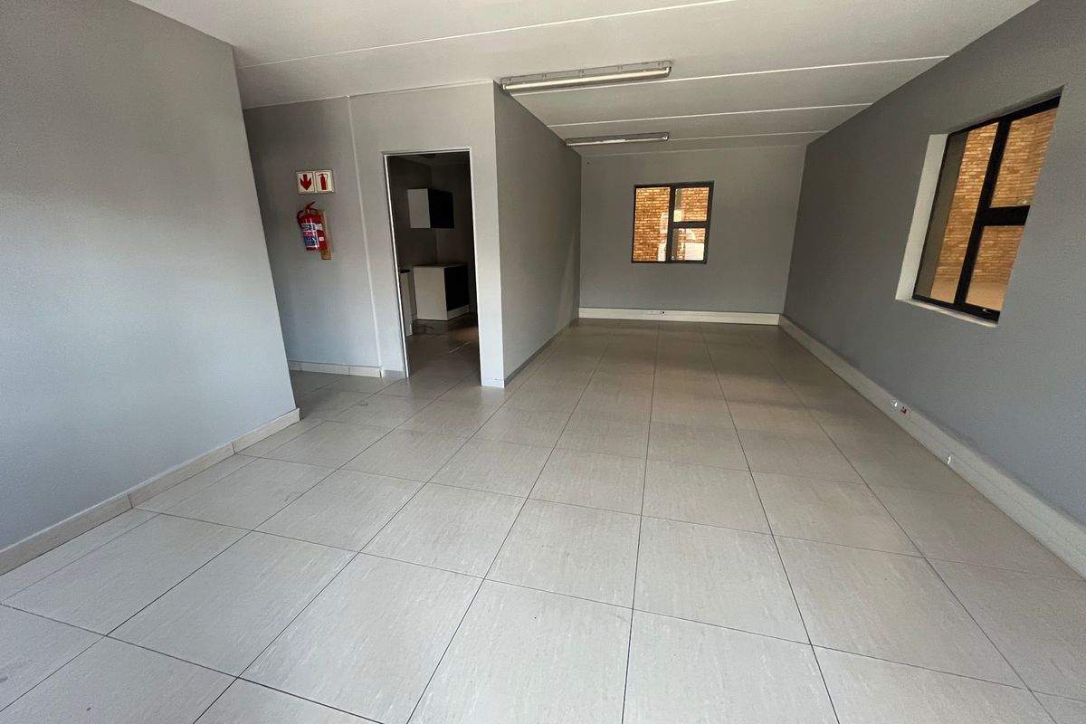 An open-plan industrial office space featuring neutral grey walls, tiled flooring, fluorescent lighting, and a wall-mounted fire extinguisher.