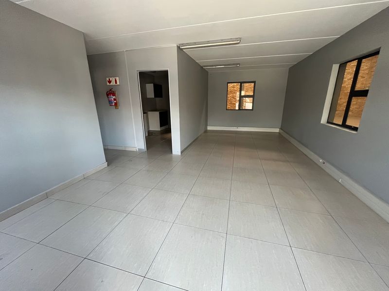 An open-plan industrial office space featuring neutral grey walls, tiled flooring, fluorescent lighting, and a wall-mounted fire extinguisher.