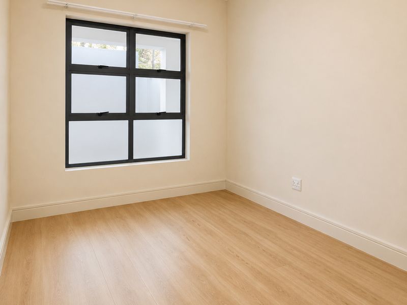 A bright, empty bedroom featuring light wood-look laminate flooring, neutral walls, and a modern black-framed window with frosted glass panes.