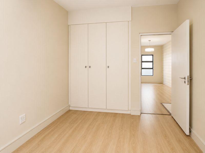 A bright, empty bedroom featuring light wood-look flooring, neutral walls, and built-in floor-to-ceiling cabinetry, with a view into the adjacent living area.