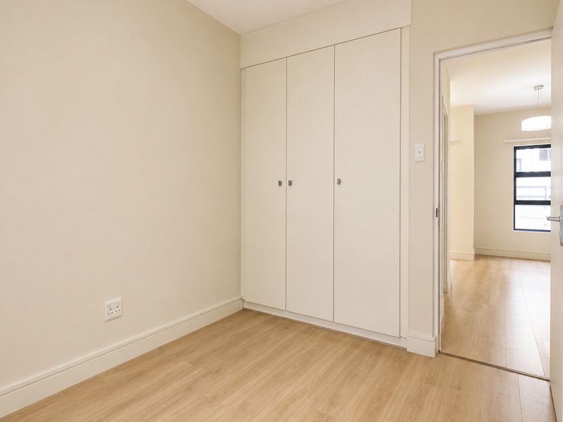 A bright, empty bedroom featuring light wood-look flooring, neutral walls, and built-in floor-to-ceiling cabinetry, with a doorway leading to an adjacent room.