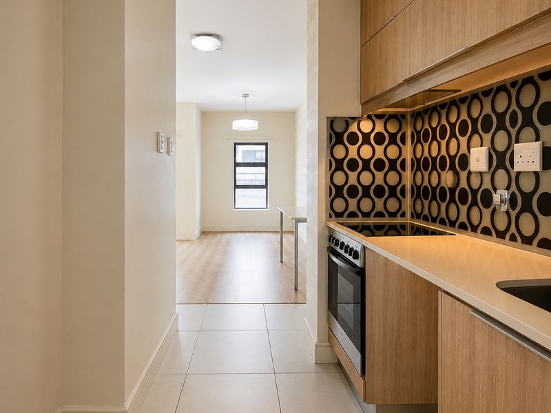 A modern galley-style kitchen featuring wood-grain cabinetry, a patterned backsplash, and integrated oven and hob, leading into an open-plan living area.