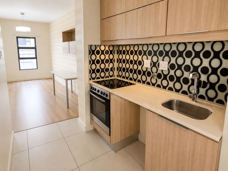 Modern open-plan kitchen featuring light wood cabinetry, integrated oven and hob, and a distinctive patterned backsplash, flowing into a bright living area.