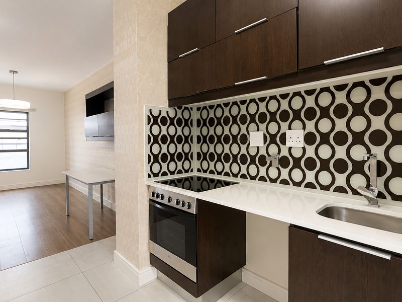 Compact kitchen featuring dark wood cabinetry, integrated oven and hob, and a patterned backsplash, flowing into an adjacent open-plan living area.