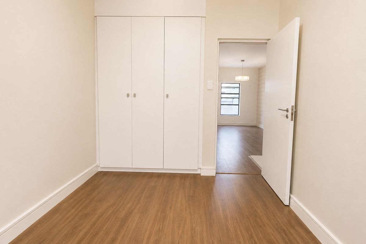 A bright, empty bedroom featuring wood-look flooring, built-in white cabinetry, and a doorway leading to an adjacent living area.