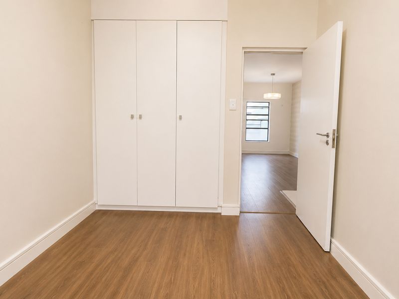 A bright, empty bedroom featuring wood-look flooring, built-in white cabinetry, and a doorway leading to an adjacent living area.