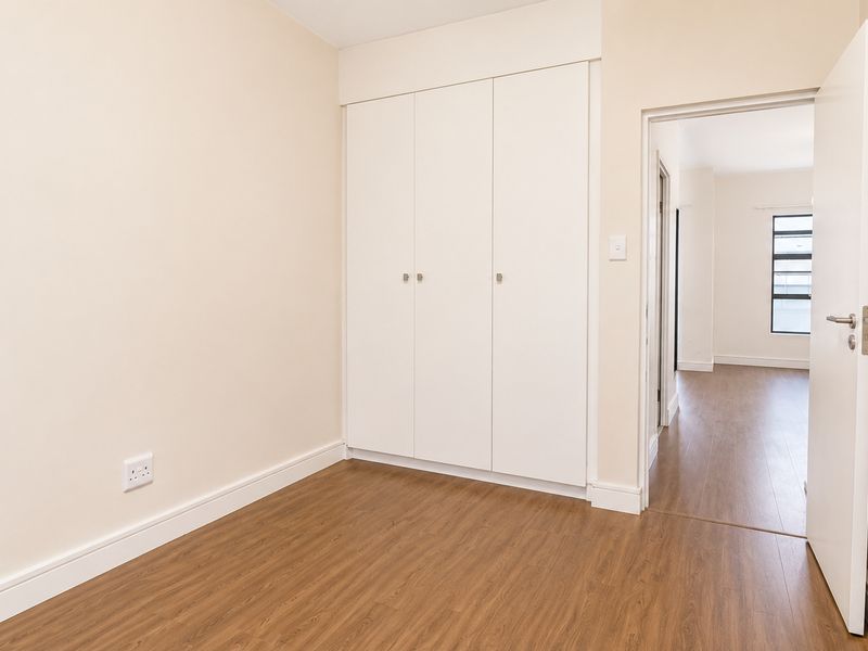 A bright, empty bedroom featuring wood-look laminate flooring and built-in white cupboards, with a doorway leading to an adjacent room.