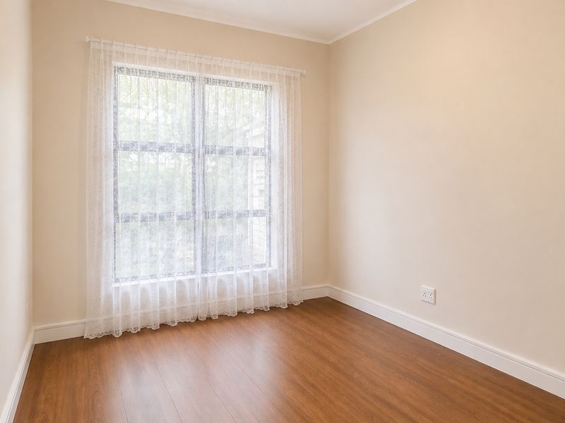 A bright, empty bedroom featuring warm-toned laminate flooring, neutral walls, and a large window with installed curtain rod.